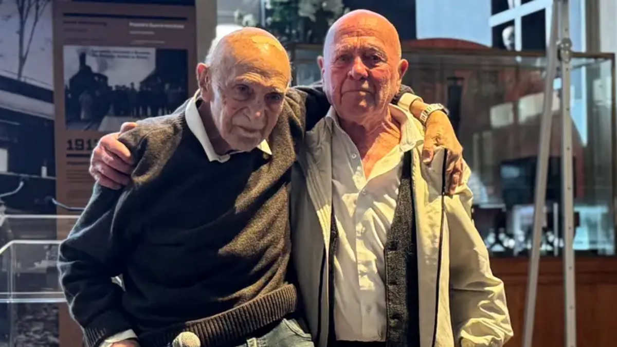 Two Holocaust survivors embracing inside a museum exhibition, standing before historical photographs and a wartime railway display.